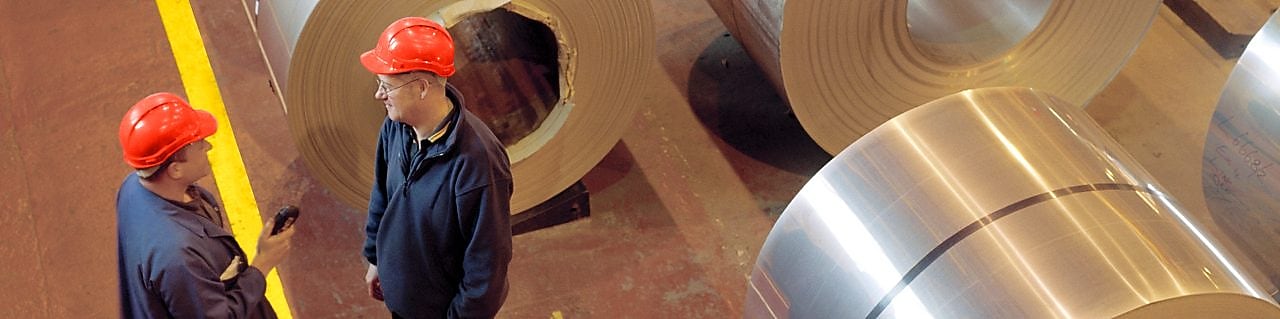 Two factory workers standing next to rolls of steel