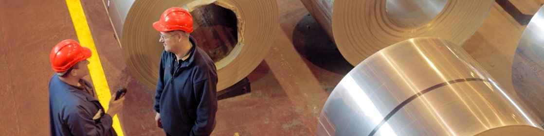 Two factory workers standing next to rolls of steel