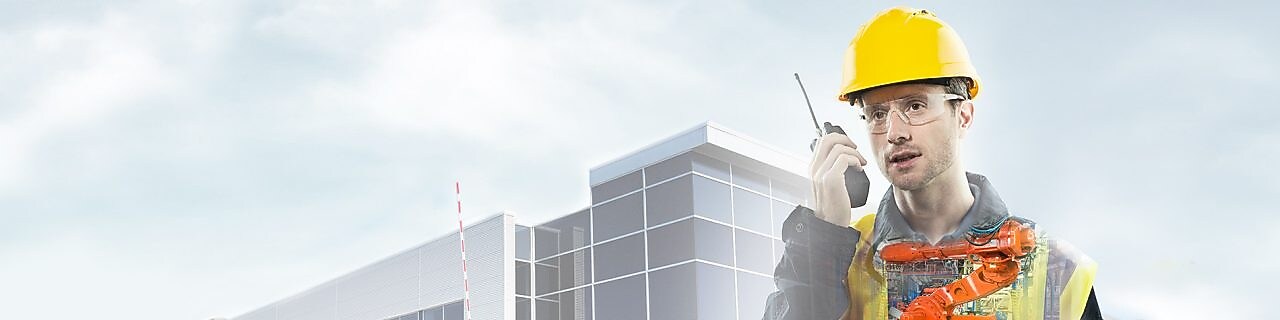 чоловік у жовтій касці говорить в рацію на тлі великої будівлі